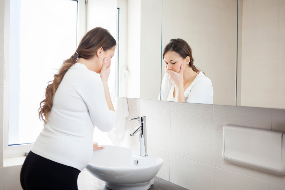Ibu hamil merasa mual dan menutup mulutnya di depan wastafel kamar mandi, mencerminkan gejala morning sickness.