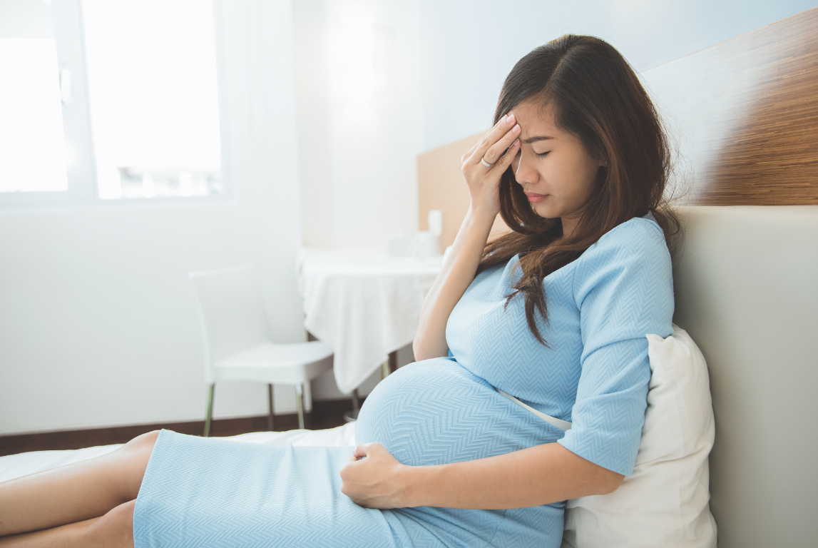 Seorang ibu hamil sedang duduk di tempat tidur sambil memegang kepala dan perutnya, tampak tidak nyaman atau pusing.