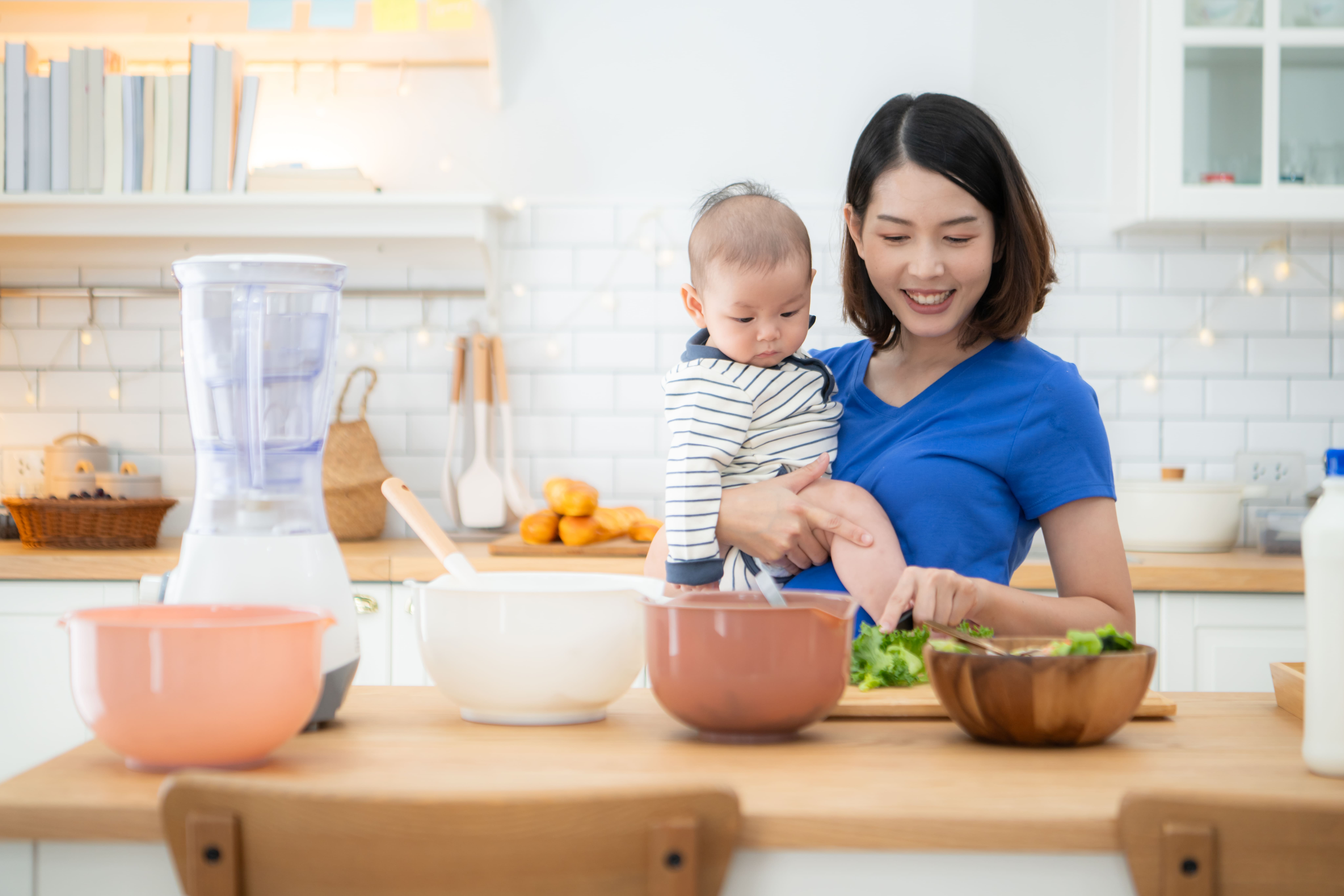 Ibu menyusui sedang menyiapkan makanan sehat sambil menggendong bayinya di dapur